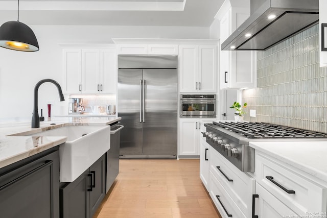 kitchen with wall chimney range hood, appliances with stainless steel finishes, hanging light fixtures, light stone counters, and white cabinets