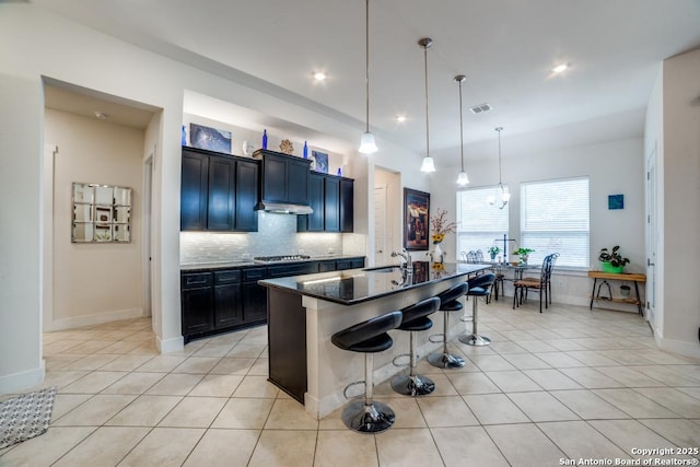 kitchen featuring a kitchen bar, stainless steel gas cooktop, light tile patterned floors, dark stone counters, and a kitchen island with sink