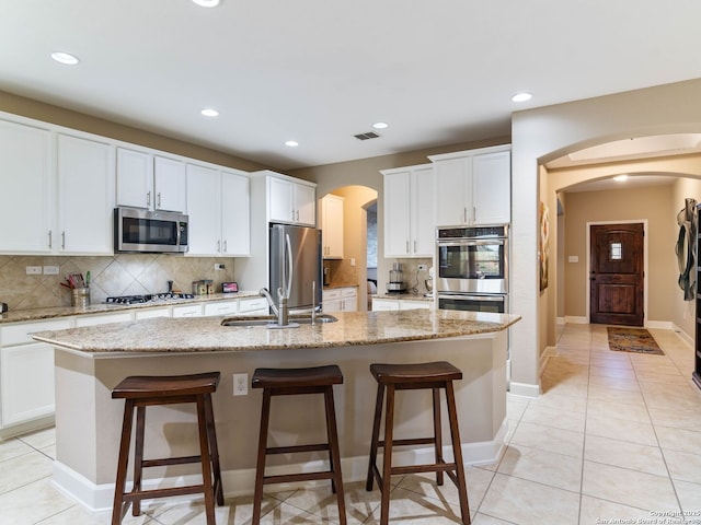 kitchen with sink, a kitchen island with sink, white cabinetry, stainless steel appliances, and light stone counters