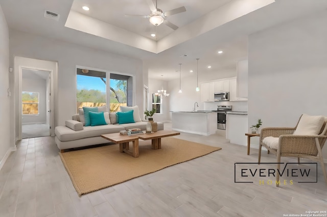 living room with sink, a tray ceiling, ceiling fan with notable chandelier, light hardwood / wood-style floors, and a high ceiling