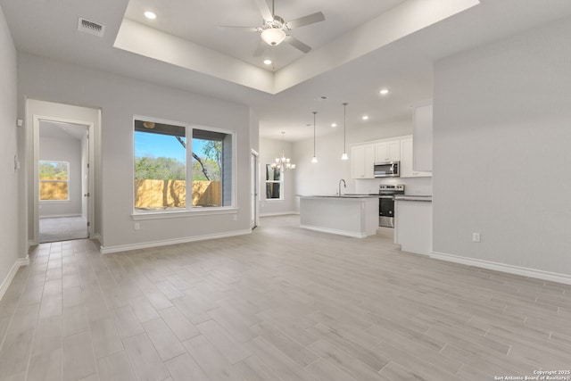 unfurnished living room featuring a tray ceiling, ceiling fan with notable chandelier, plenty of natural light, and light wood-type flooring