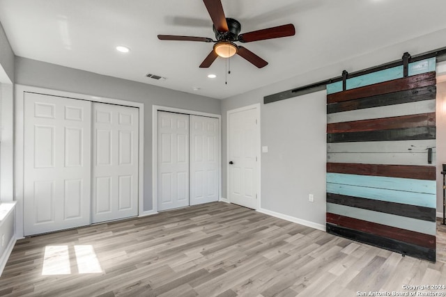 unfurnished bedroom with a barn door, two closets, ceiling fan, and light hardwood / wood-style flooring