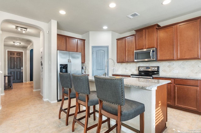 kitchen featuring an island with sink, sink, backsplash, stainless steel appliances, and light stone countertops