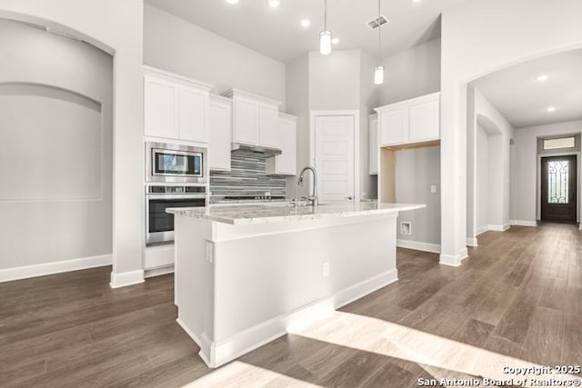 kitchen with white cabinets, hanging light fixtures, a kitchen island with sink, stainless steel appliances, and light stone countertops