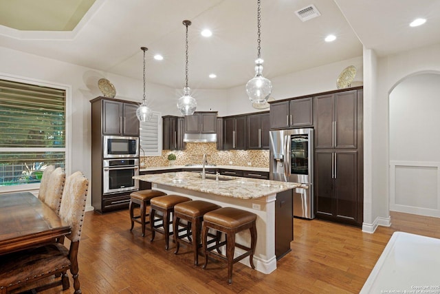 kitchen featuring appliances with stainless steel finishes, pendant lighting, light stone countertops, a kitchen island with sink, and backsplash