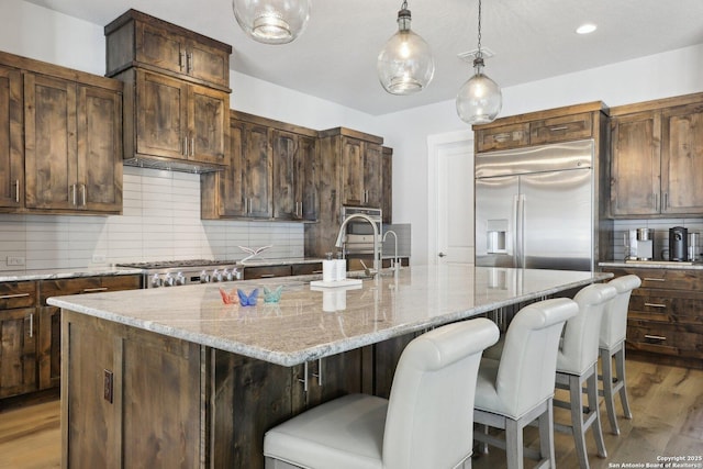 kitchen with appliances with stainless steel finishes, light stone countertops, a kitchen island with sink, and backsplash