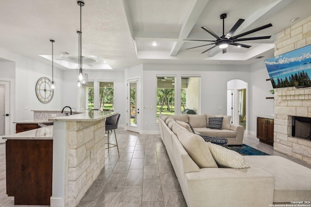 living room featuring ceiling fan, arched walkways, coffered ceiling, and a stone fireplace