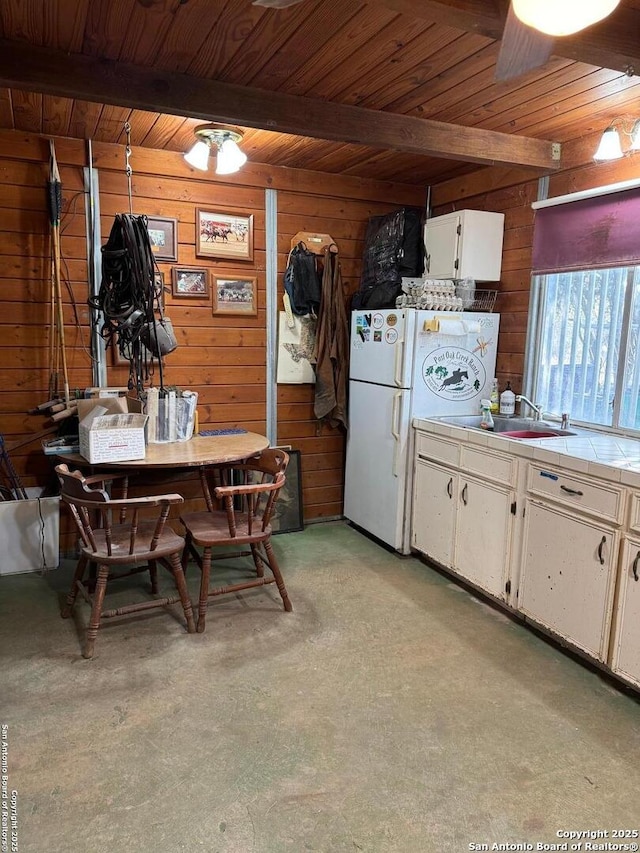 kitchen with wooden walls, white cabinets, tile countertops, a sink, and beam ceiling