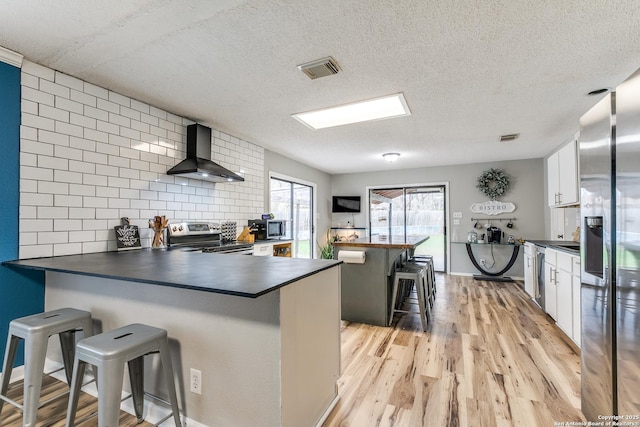 kitchen with stainless steel appliances, wall chimney exhaust hood, white cabinets, and a kitchen bar