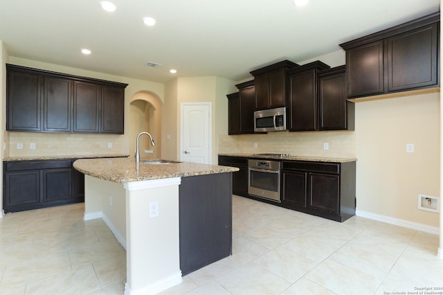 kitchen featuring sink, dark brown cabinetry, light stone counters, stainless steel appliances, and a center island with sink