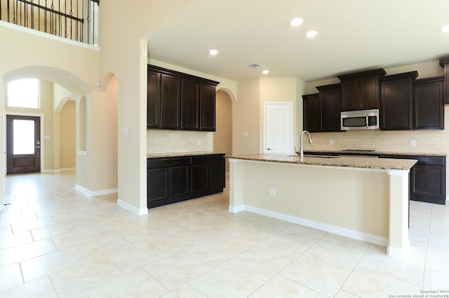 kitchen featuring sink, dark brown cabinets, light stone counters, tasteful backsplash, and a center island with sink