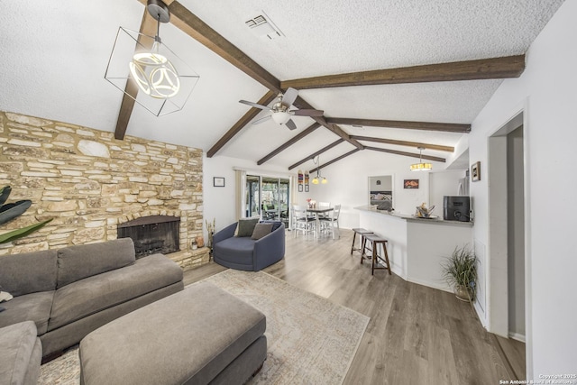 living room with a stone fireplace, vaulted ceiling with beams, light hardwood / wood-style flooring, a textured ceiling, and ceiling fan