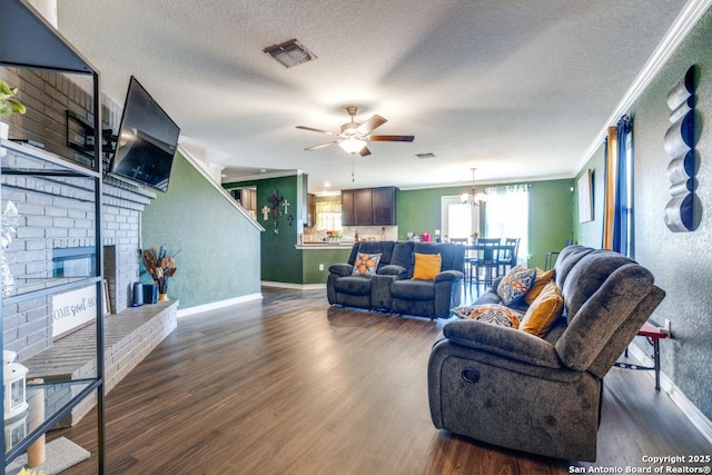 living room featuring dark wood-type flooring, a brick fireplace, a textured ceiling, ornamental molding, and ceiling fan with notable chandelier