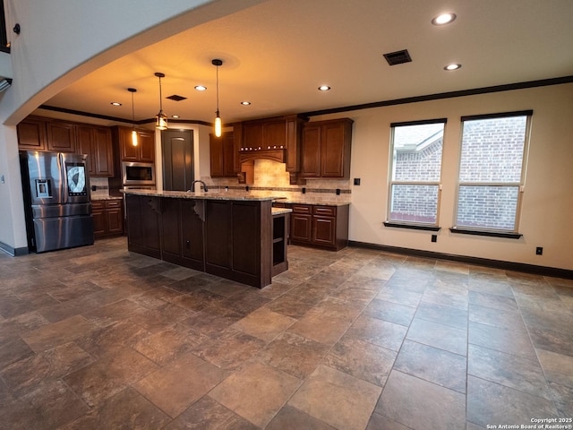 kitchen featuring appliances with stainless steel finishes, hanging light fixtures, a kitchen island with sink, light stone counters, and crown molding