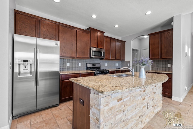 kitchen featuring sink, light stone counters, stainless steel appliances, a kitchen island with sink, and decorative backsplash