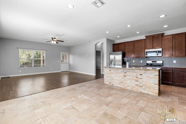 kitchen featuring ceiling fan, stainless steel appliances, light stone countertops, an island with sink, and decorative backsplash