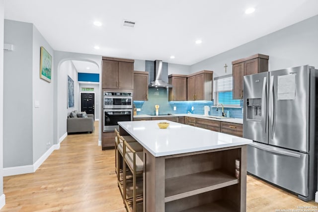 kitchen featuring backsplash, a center island, stainless steel appliances, light wood-type flooring, and wall chimney exhaust hood