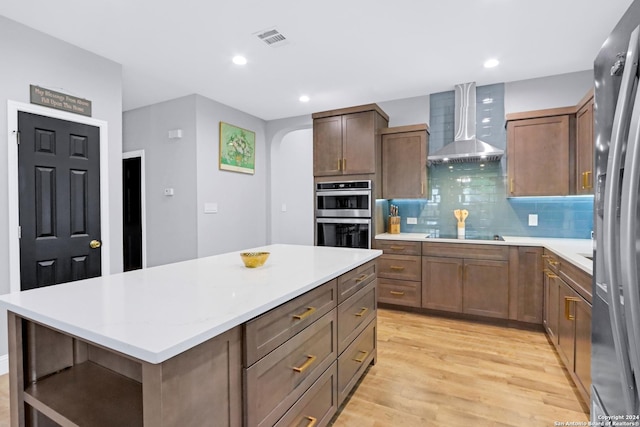 kitchen with a kitchen island, appliances with stainless steel finishes, tasteful backsplash, wall chimney range hood, and light wood-type flooring