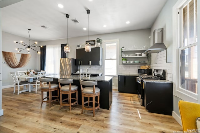 kitchen with wall chimney exhaust hood, a breakfast bar, a kitchen island with sink, stainless steel appliances, and light hardwood / wood-style floors