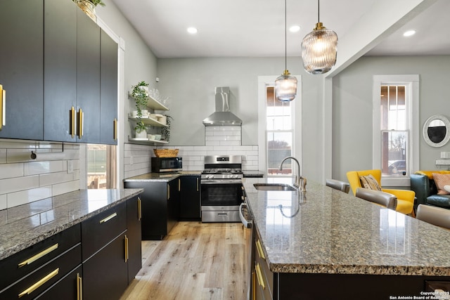 kitchen with stainless steel range with gas stovetop, an island with sink, hanging light fixtures, and wall chimney range hood