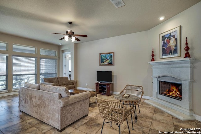 living room with a warm lit fireplace, baseboards, visible vents, a ceiling fan, and a textured ceiling