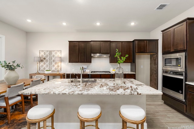 kitchen featuring sink, stainless steel appliances, dark brown cabinetry, light stone countertops, and an island with sink