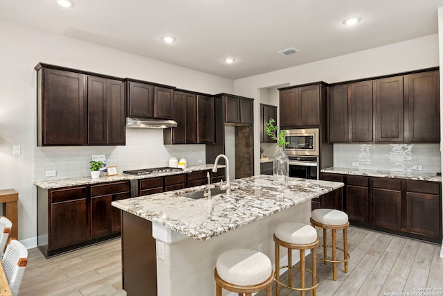 kitchen with sink, light wood-type flooring, an island with sink, stainless steel appliances, and light stone countertops