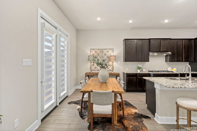 kitchen featuring stainless steel gas stovetop, sink, decorative backsplash, dark brown cabinetry, and light stone countertops