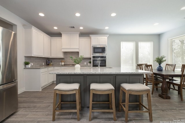 kitchen featuring sink, appliances with stainless steel finishes, white cabinetry, light stone counters, and a center island with sink