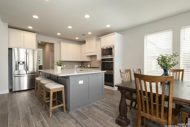 kitchen with appliances with stainless steel finishes, white cabinetry, backsplash, a kitchen island with sink, and light stone counters