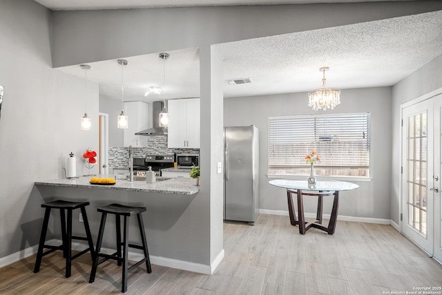 kitchen with wall chimney range hood, stainless steel appliances, white cabinets, a kitchen bar, and decorative light fixtures