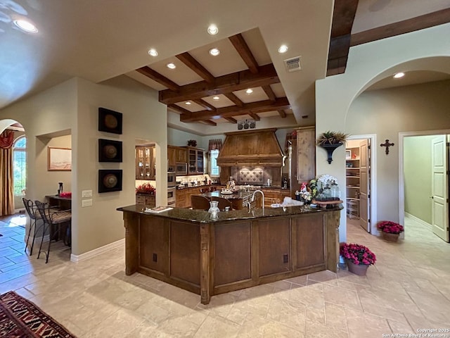 kitchen with premium range hood, coffered ceiling, stainless steel microwave, dark stone counters, and beamed ceiling