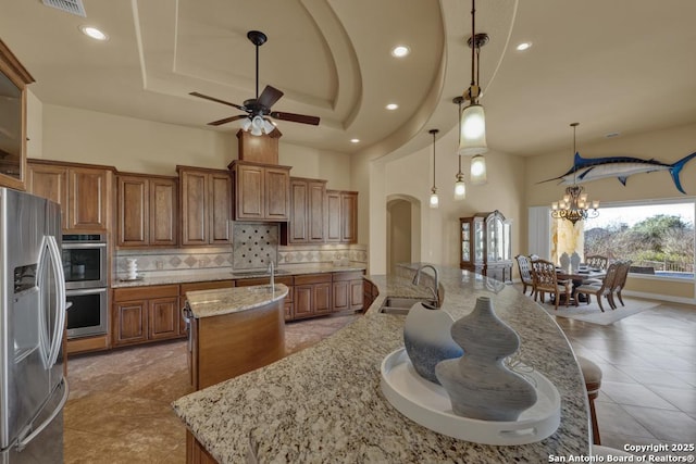 kitchen with sink, appliances with stainless steel finishes, a kitchen island with sink, light stone counters, and decorative light fixtures