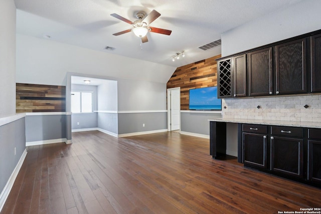 kitchen featuring lofted ceiling, dark wood-type flooring, dark brown cabinets, wooden walls, and light stone counters