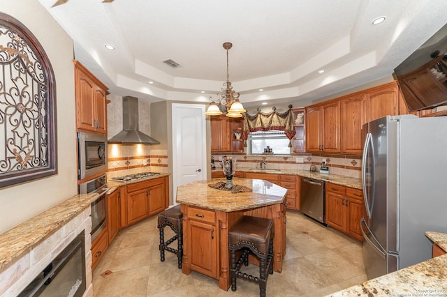 kitchen with wall chimney range hood, appliances with stainless steel finishes, a kitchen breakfast bar, a center island, and a raised ceiling