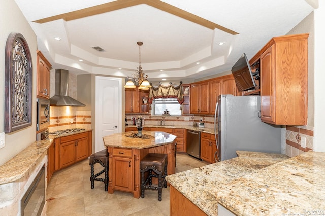 kitchen with stainless steel appliances, a center island, wall chimney range hood, and a tray ceiling