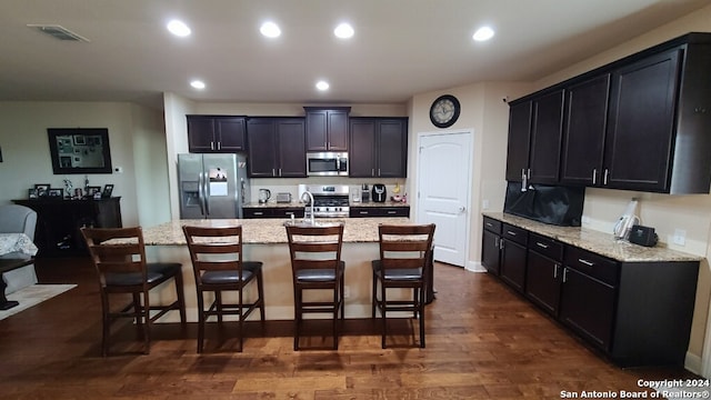 kitchen with light stone counters, a center island with sink, appliances with stainless steel finishes, dark hardwood / wood-style floors, and a kitchen breakfast bar