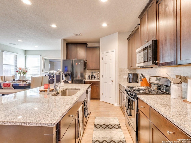 kitchen featuring sink, appliances with stainless steel finishes, tasteful backsplash, light stone countertops, and an island with sink