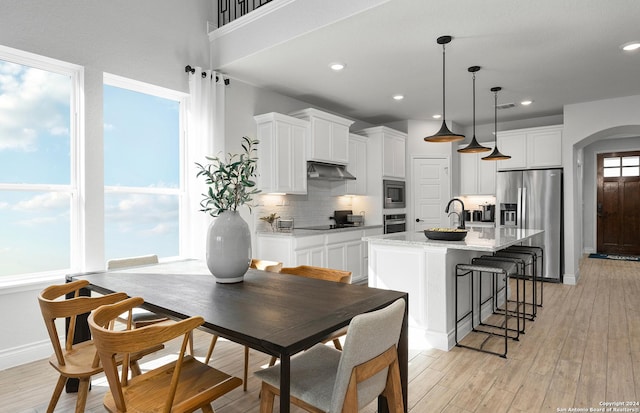 kitchen with white cabinetry, light stone counters, hanging light fixtures, appliances with stainless steel finishes, and a kitchen island with sink