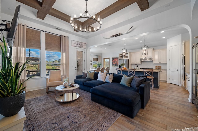 living room with sink, a towering ceiling, beamed ceiling, light hardwood / wood-style floors, and a chandelier