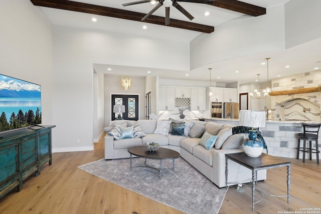 living room with beamed ceiling, a high ceiling, ceiling fan with notable chandelier, and light hardwood / wood-style flooring