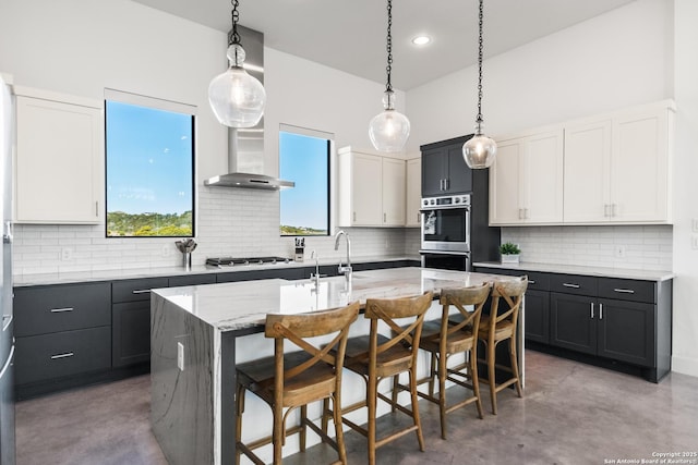 kitchen featuring double oven, white cabinets, dark cabinetry, wall chimney exhaust hood, and a center island with sink