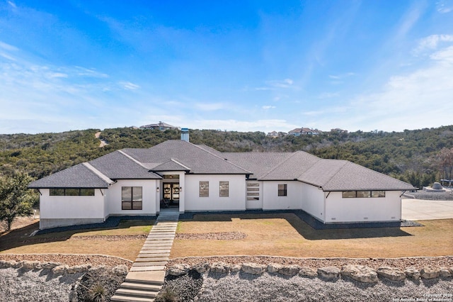 view of front facade with a forest view, a shingled roof, a front lawn, and stucco siding