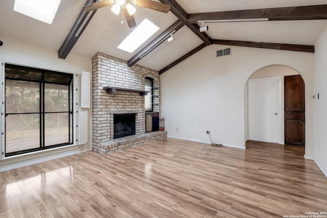 unfurnished living room with vaulted ceiling with skylight, ceiling fan, a brick fireplace, a textured ceiling, and light wood-type flooring