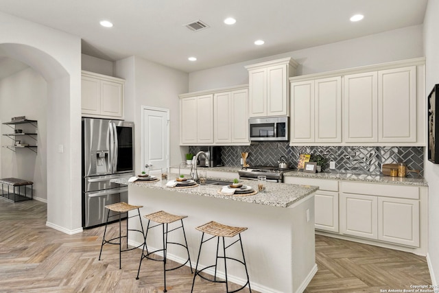 kitchen with light stone counters, stainless steel appliances, a center island with sink, and light parquet floors