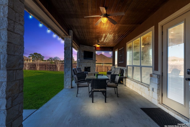 patio terrace at dusk featuring ceiling fan, a lawn, and an outdoor stone fireplace