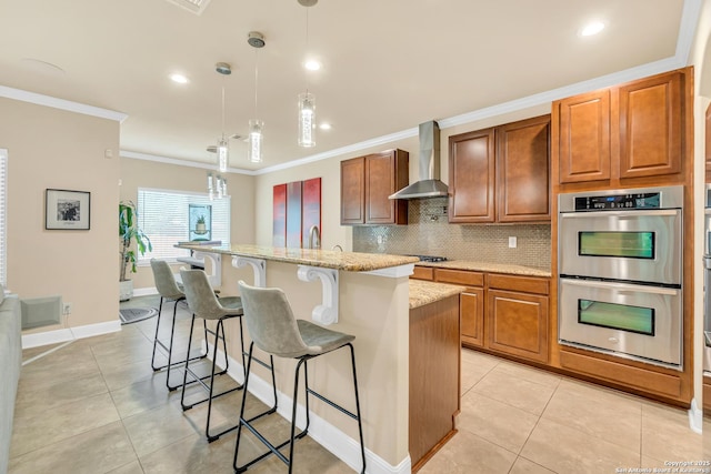 kitchen featuring light stone counters, decorative light fixtures, a center island, double oven, and wall chimney range hood