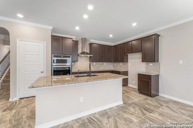 kitchen with sink, stainless steel double oven, light stone countertops, a center island with sink, and wall chimney exhaust hood