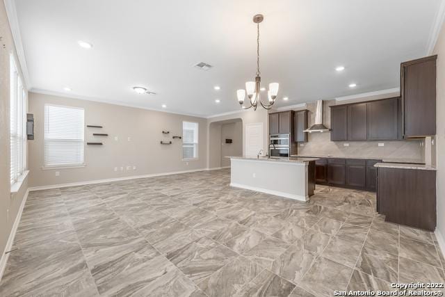 kitchen with decorative backsplash, crown molding, an island with sink, and wall chimney exhaust hood