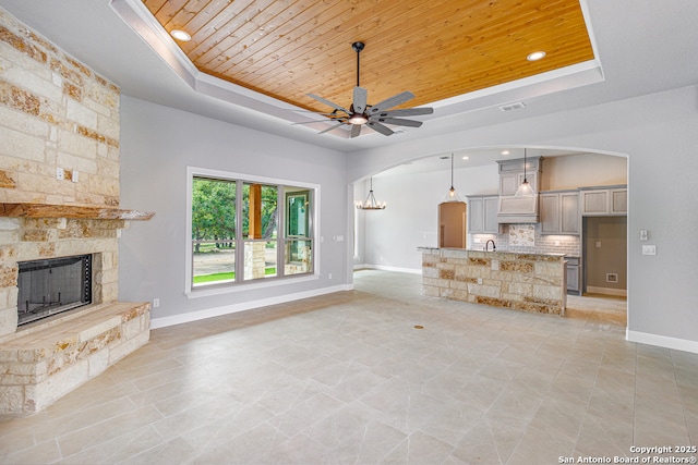 unfurnished living room with a stone fireplace, ceiling fan with notable chandelier, wooden ceiling, and a tray ceiling